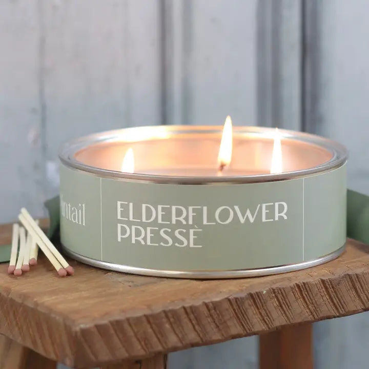 Candle in a tin labeled 'Elderflower Presse' on a wooden surface with matches beside it.