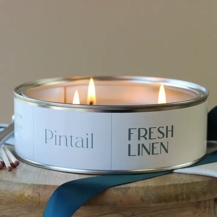 Candle in a tin labeled 'Pintail Fresh Linen' with a ribbon and matches on a wooden surface.