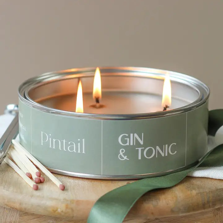 Candle in a green tin labeled 'Pintail Gin & Tonic' with matches on a wooden surface.