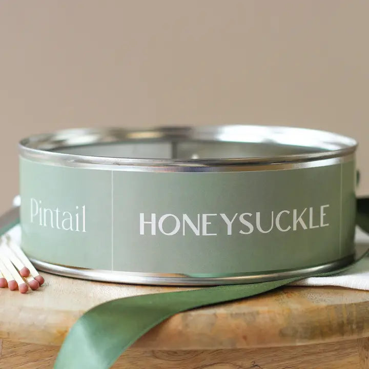 Green candle tin labeled 'Honeysuckle' on a wooden surface with a beige background