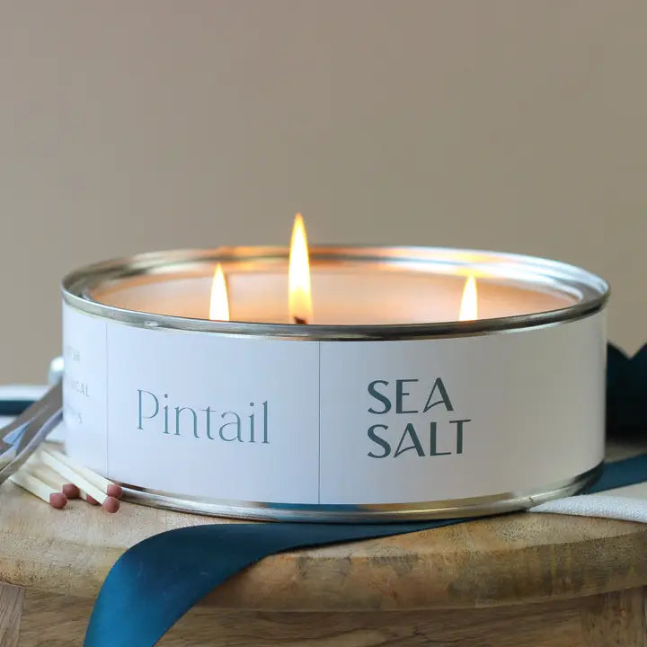 Candle in a tin labeled 'Pintail Sea Salt' on a wooden surface with matches.