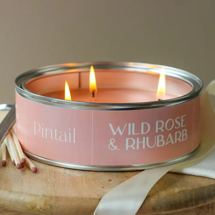 Candle in a pink tin labeled 'Wild Rose & Rhubarb' with matches and ribbon on a wooden surface.