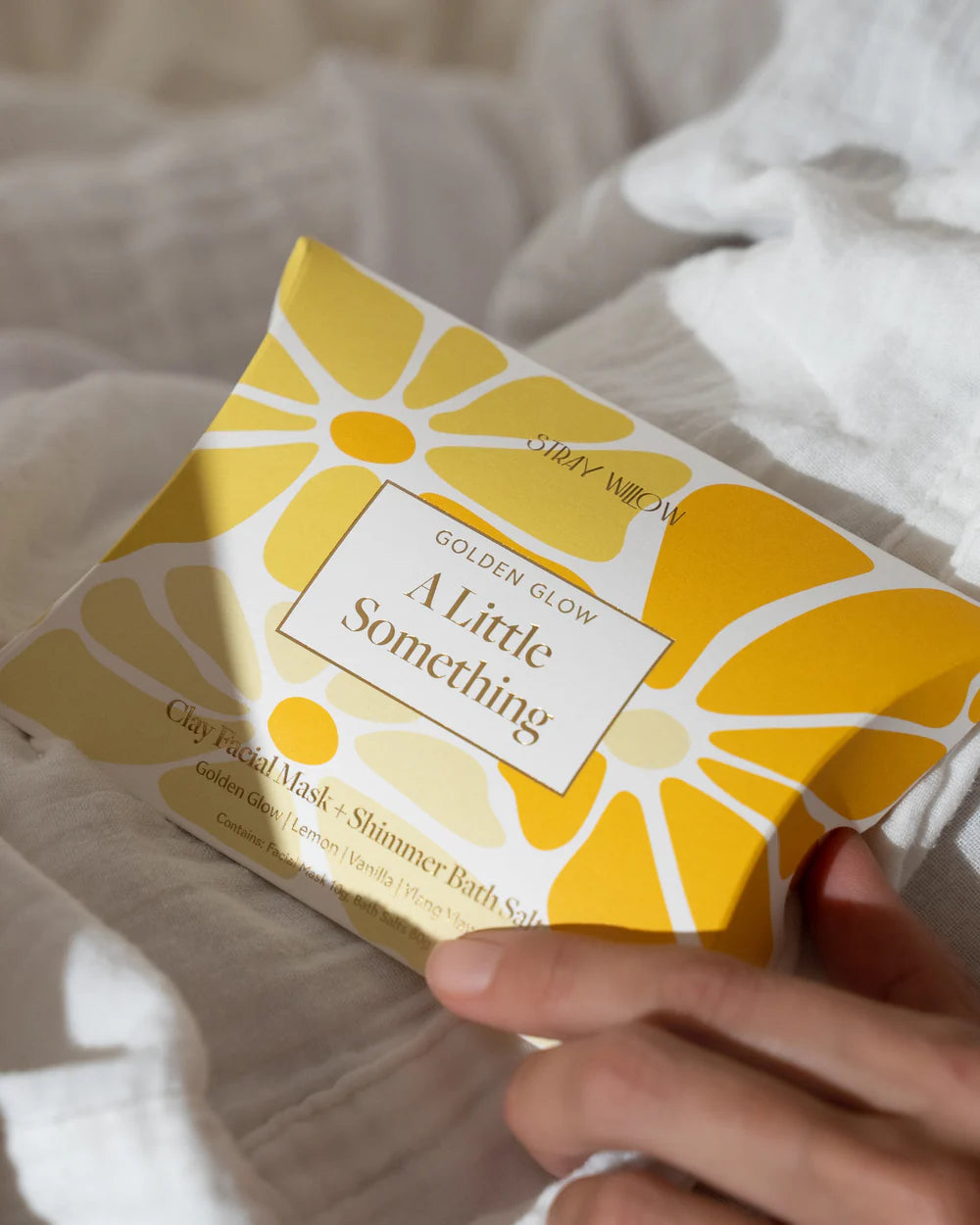 Yellow packaging of 'A Little Something' product held by a hand on a white fabric background