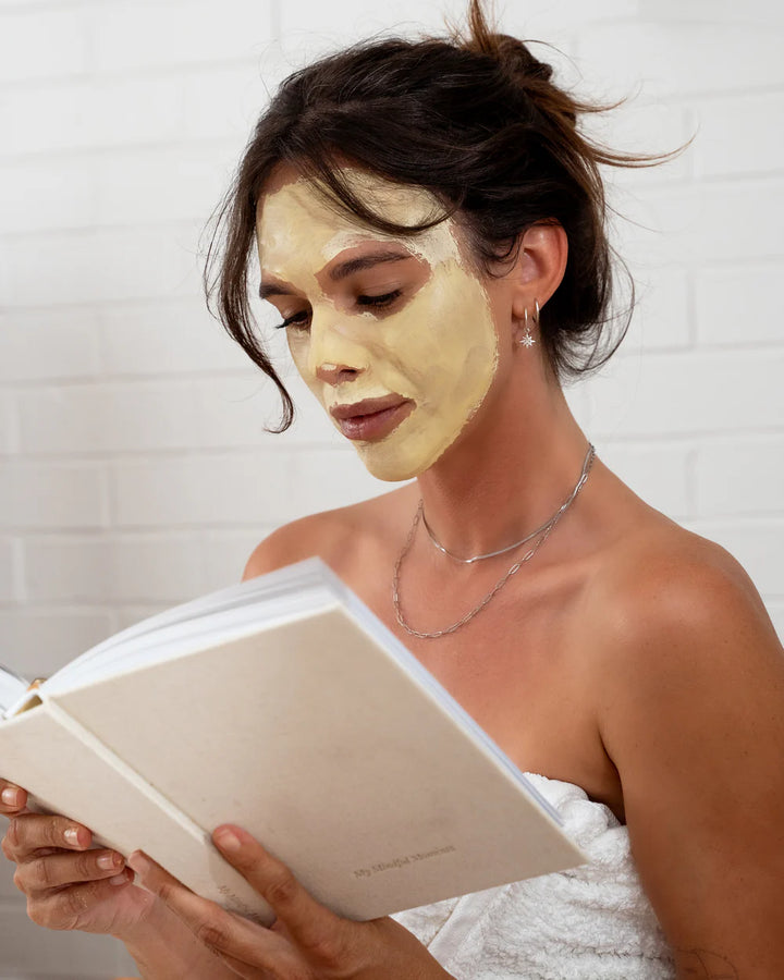 Woman with a facial mask reading a book in a bathroom.