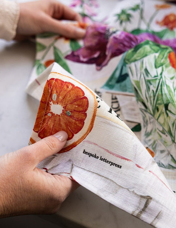 Hand holding a linen tea towel with a grapefruit illustration and 'bespoke letterpress' text, against a floral-patterned fabric background.