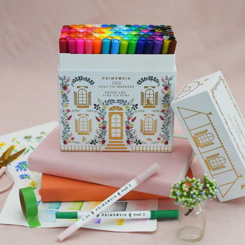 Box of Primrosia dual-tip markers with a pink notebook and green pen on a light pink background