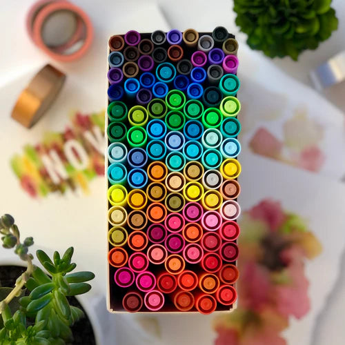 Box of colorful markers on a blurred background with plants and stationery items.