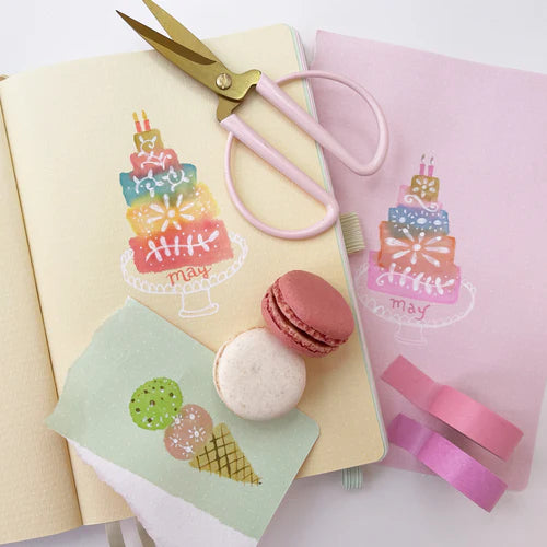 Notebook with cake illustration, pink scissors, and macaron decorations on a white background