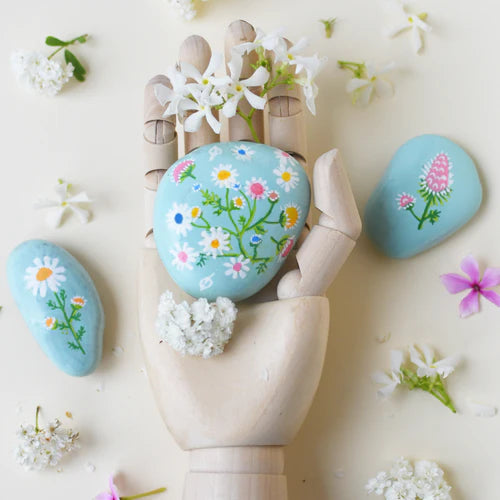 Decorative stones with floral designs held by a wooden hand, surrounded by flowers on a light background