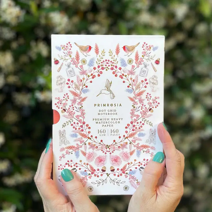 Person holding a Primrosia notebook with floral design against a blurred natural background