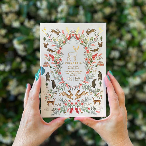 Person holding a notebook called Primrosa watercolor paper with nature-themed design against a blurred green and white floral background.