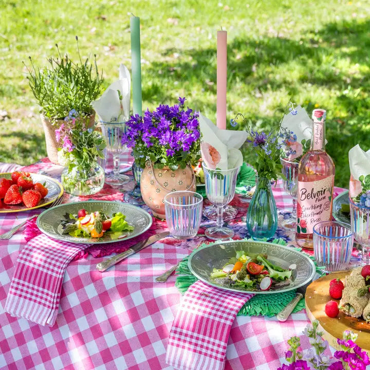 Outdoor table setting with pink checkered tablecloth, plates of food, and floral decorations.