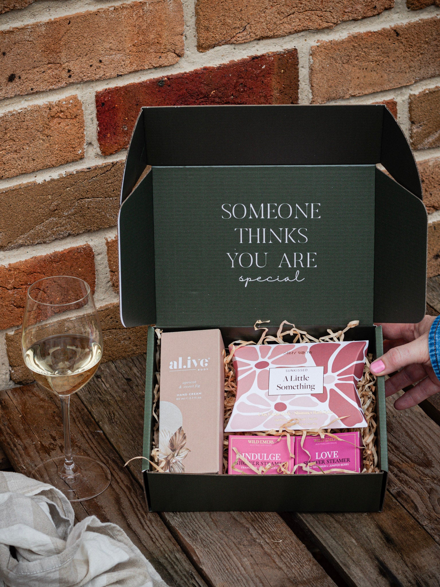 Gift box with products and a glass of wine on a wooden surface against a brick wall.