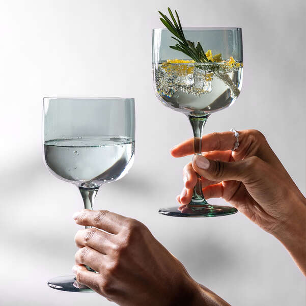 Two hands holding wine glasses with one containing a cocktail garnished with a sprig of rosemary and lemon peel.