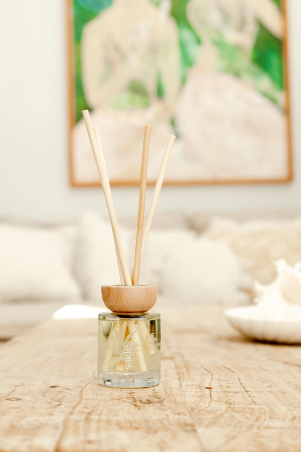 Reed diffuser with wooden holder on a wooden surface, blurred artwork in the background