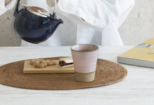 Ceramic cup on a woven placemat with a teapot and books in the background.