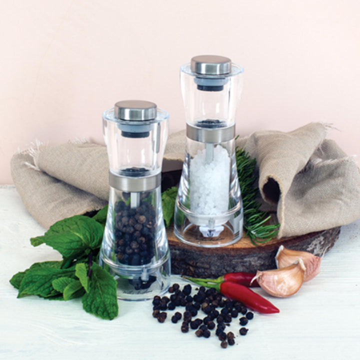Two glass pepper grinders with black and white peppercorns on a wooden board with herbs and spices.