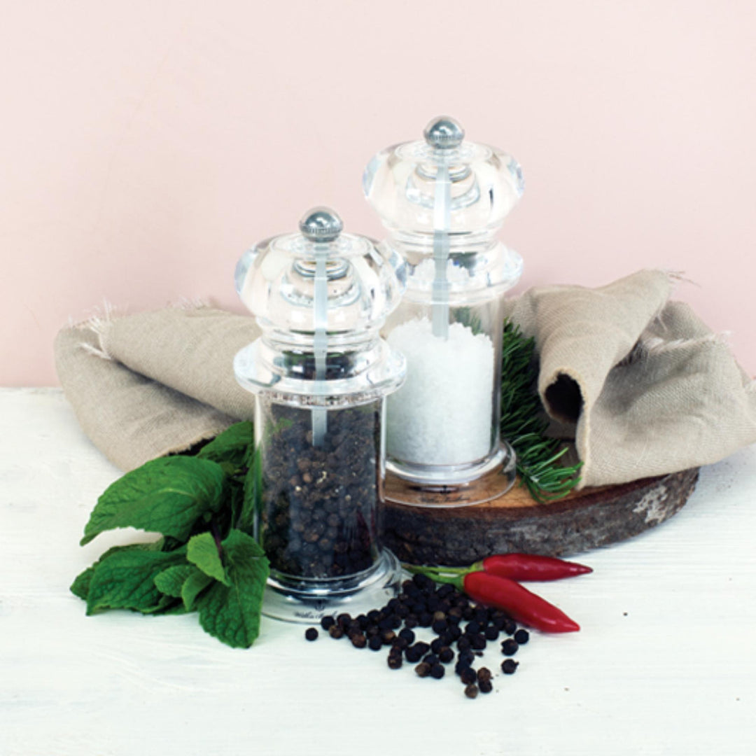 Clear pepper grinder and salt shaker with black peppercorns and red peppers on a white surface.
