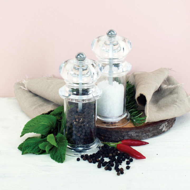 Clear pepper grinder and salt shaker with black peppercorns and red peppers on a white surface.