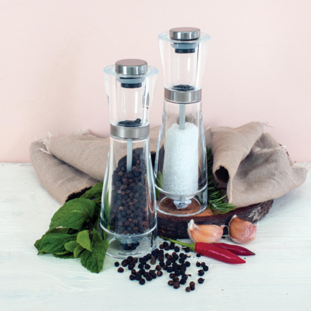 Glass salt and pepper shakers with black and white contents on a wooden surface with herbs and spices.