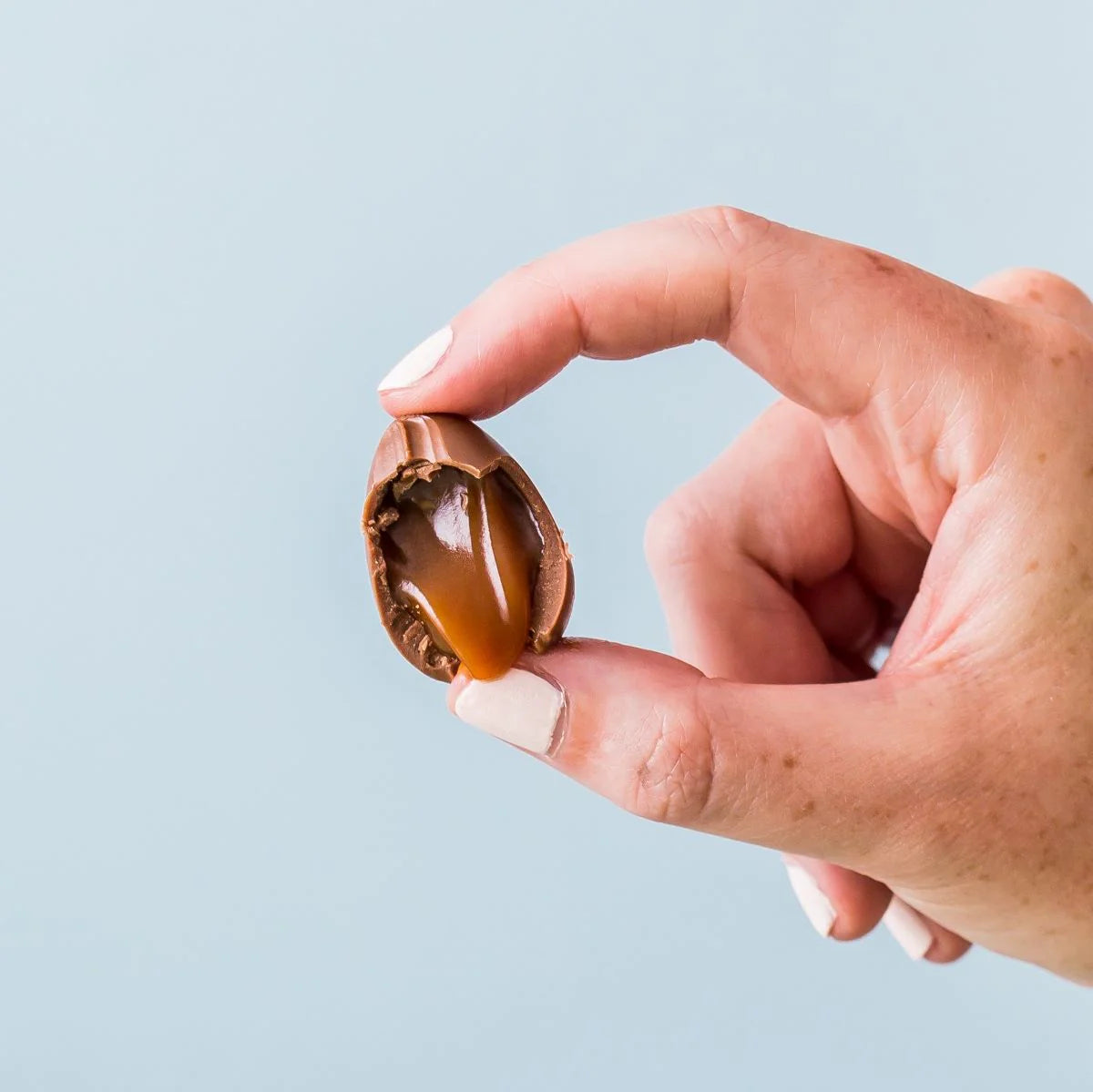 Hand holding a chocolate shell with a caramel center against a light blue background