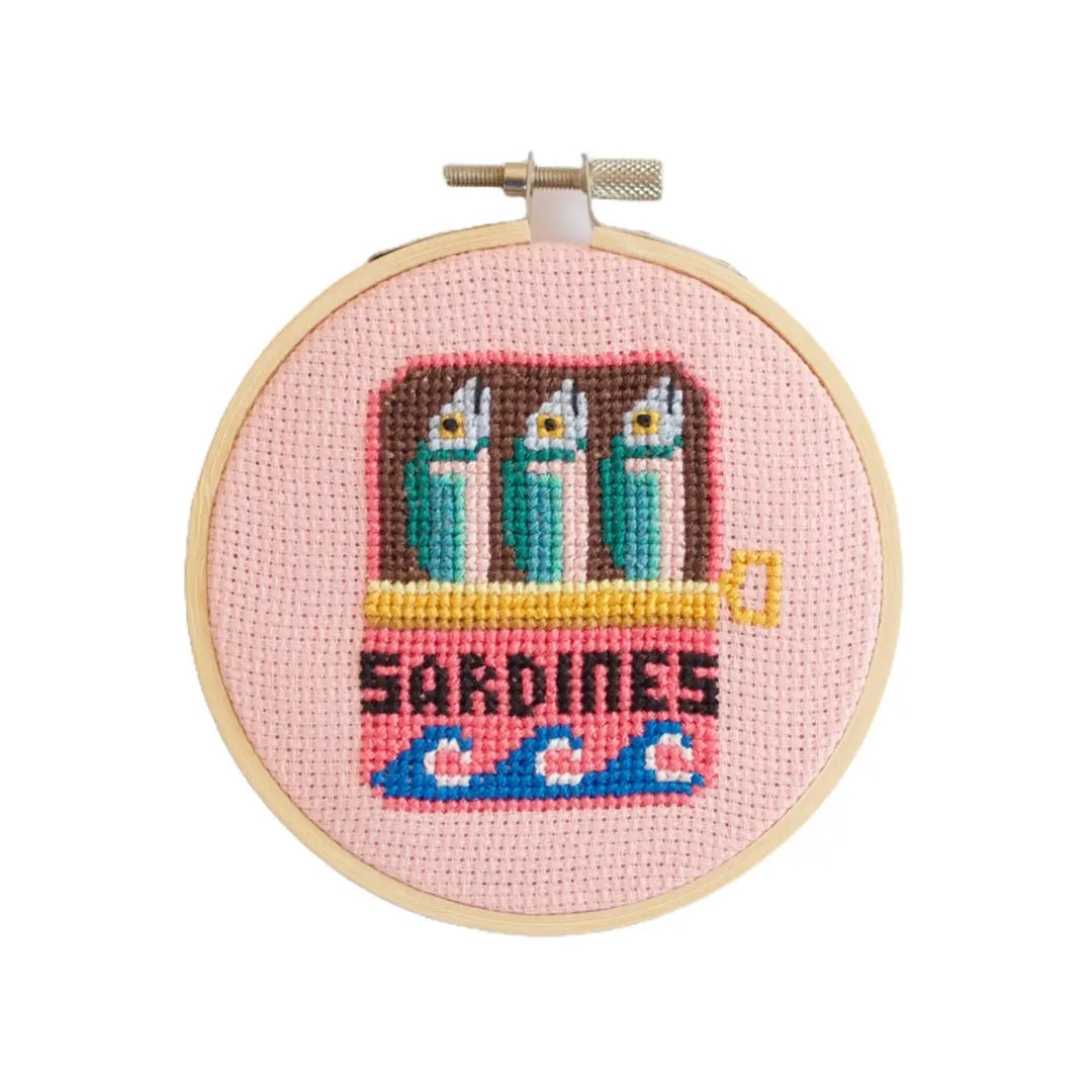 Cross-stitched design of sardines in a can on a pink background with 'Sardines' text.