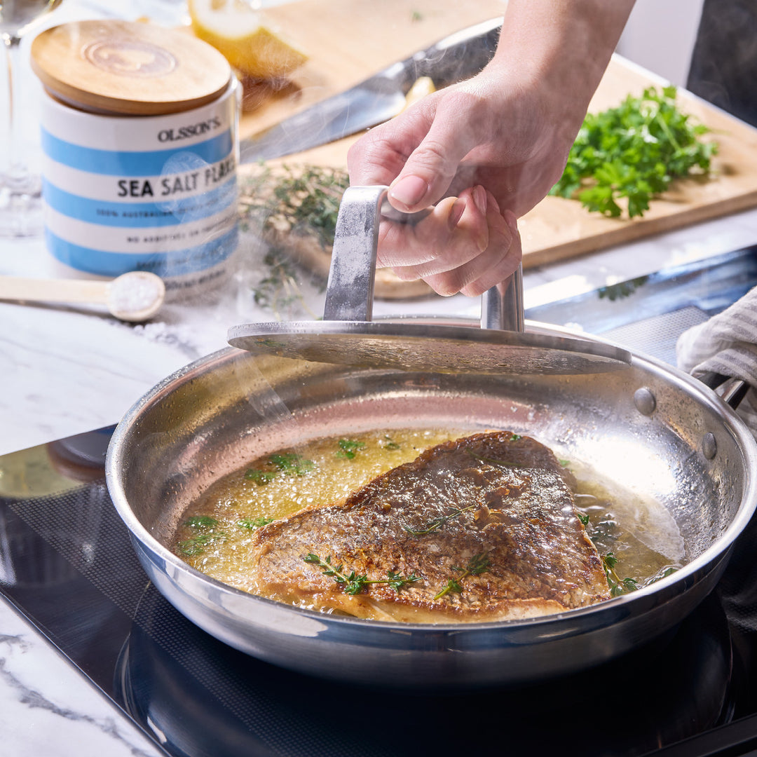 Person cooking fish in a pan on a stove with a container of sea salt in the background.