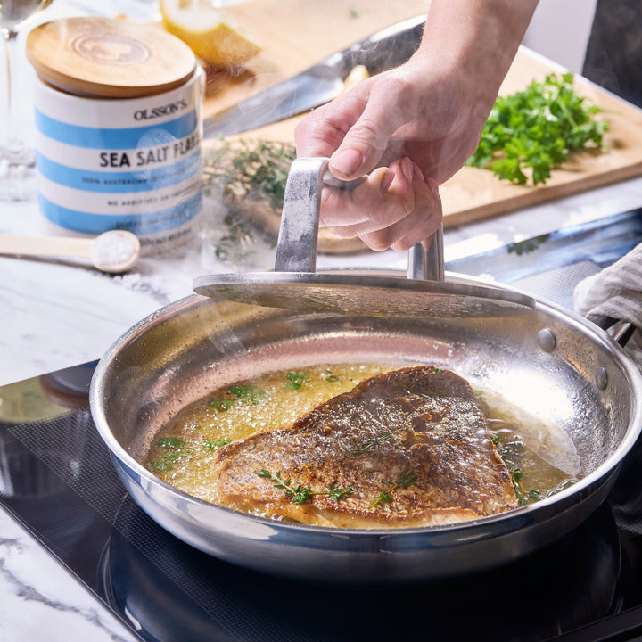 Person cooking fish in a pan on a stove with a container of sea salt in the background.