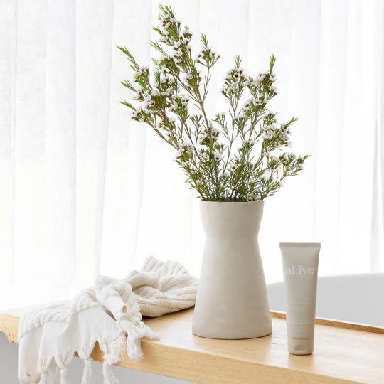White vase with greenery on a wooden surface next to a white towel and bottle.
