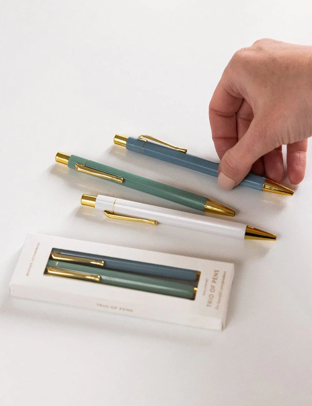 Set of pens with gold accents on a white surface, with a hand reaching for one pen.