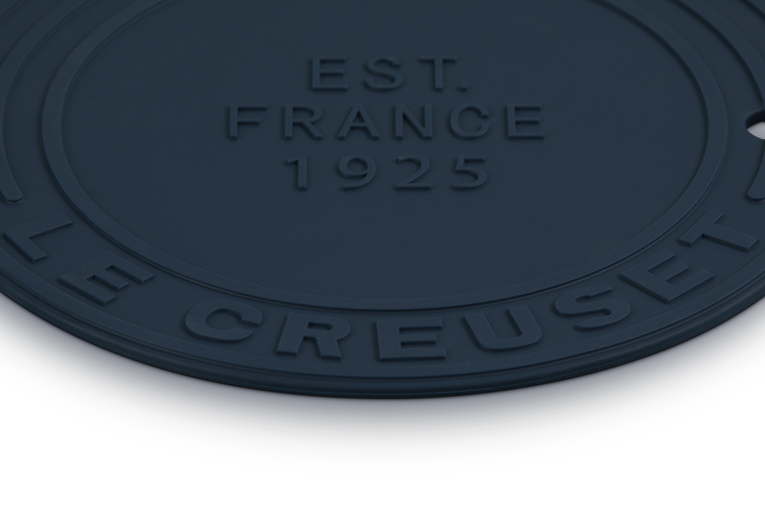 Round navy mat with 'Le Creuset' branding and 'EST. FRANCE 1925' text on a white background