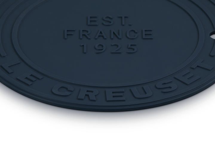 Round navy mat with 'Le Creuset' branding and 'EST. FRANCE 1925' text on a white background