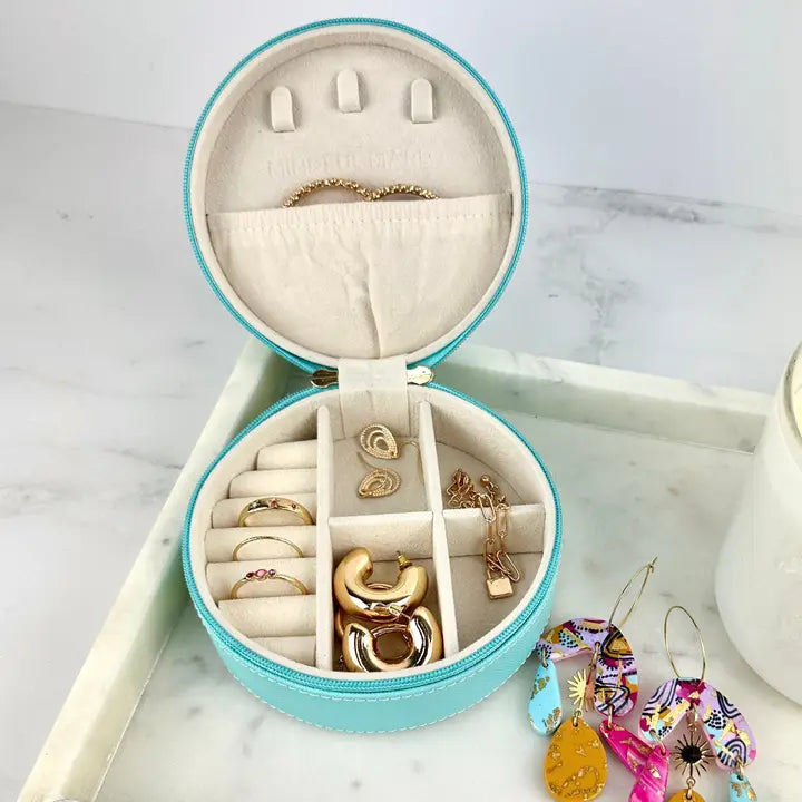 An open round turquoise jewelry organizer box with cream velvet lining, holding several gold rings and earrings, placed on a white marble surface with additional colorful earrings nearby.