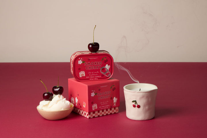 Cherry-themed product set including a box, candle in a vessel and a candle in a tin, and cup on a red surface.