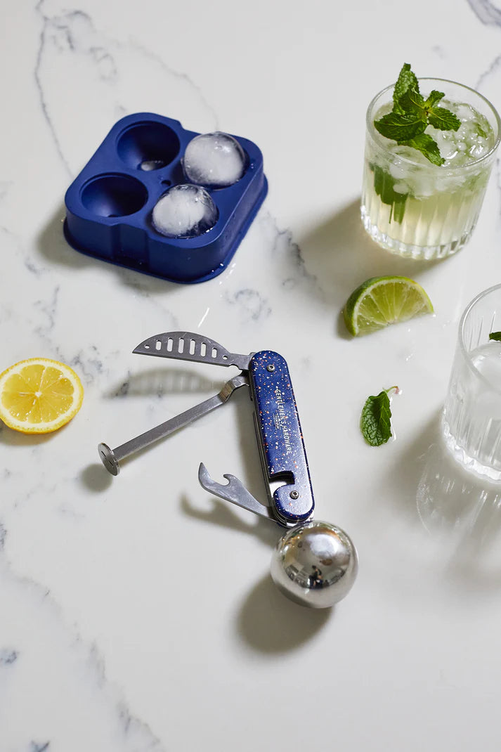 Blue ice cube tray with ice cubes, cocktail shaker, lemon, lime, and glasses on a marble surface.