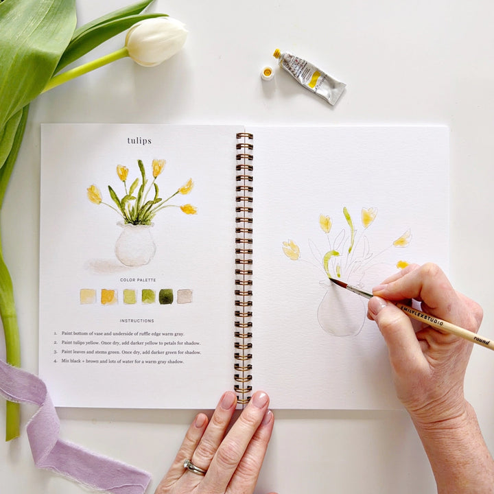 Person painting tulips on a sketchbook with a bottle of water and a tulip flower nearby.