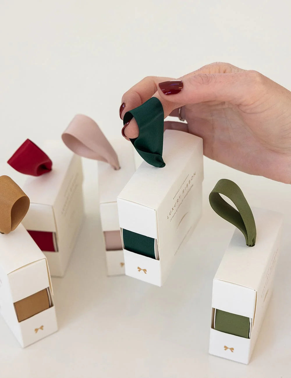 Hand holding a green ribbon over a white box with a bow, surrounded by other boxes with different colored ribbons.