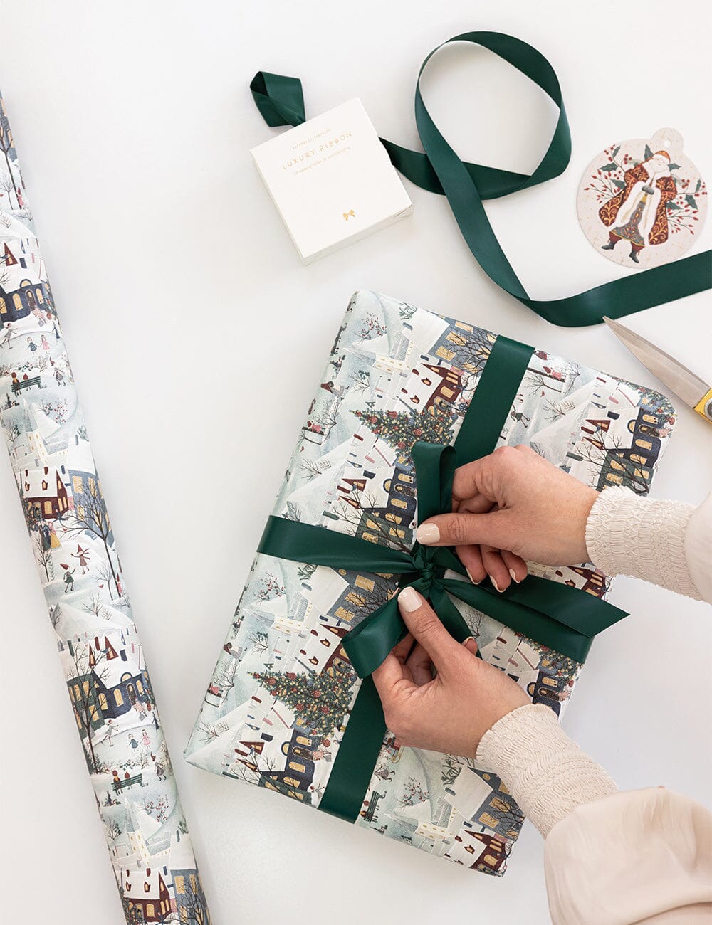 Person wrapping a gift with festive paper and green ribbon on a white surface