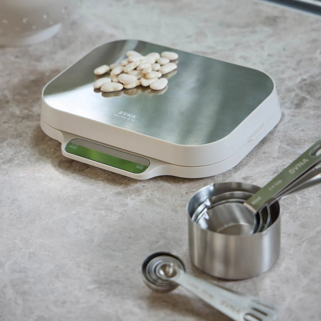 Kitchen scale with beans on a countertop, accompanied by measuring cups and spoons.