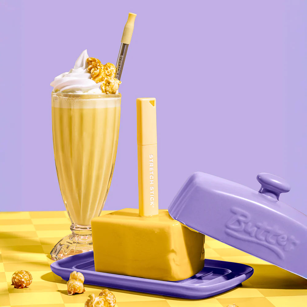 A slab of butter in a purple butter keeper, with a banana milkshake in a glass with a stainless steel straw, on a yellow surface &amp; purple background.