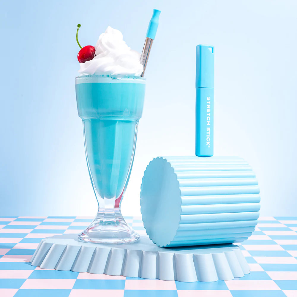 A blue coloured cocktail with a cherry on top with cream and a reusable stainless steel straw, on a blue checked surface and blue background
