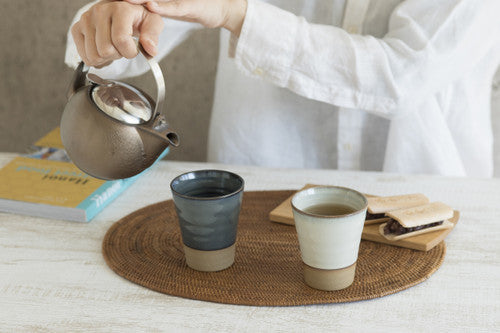 Person pouring tea from a teapot into two cups on a table with a woven mat.