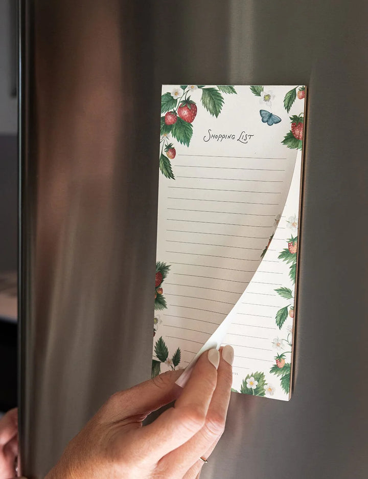 Hand holding a notepad with a floral design attached to a refrigerator.