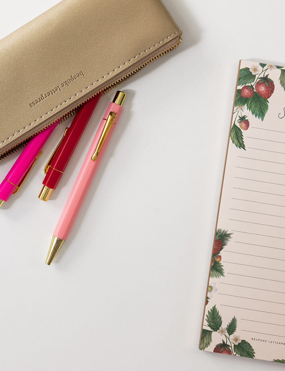 Pink pen with gold accents, red pen, beige wallet, and floral notepad on a white surface
