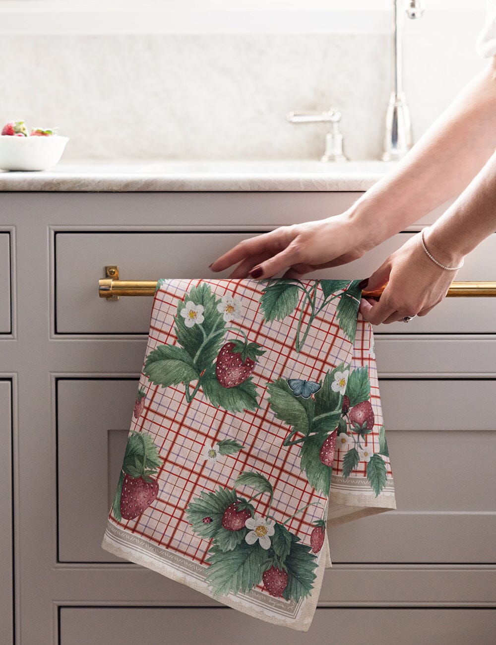 Person hanging a floral and strawberry patterned towel on a kitchen cabinet handle.