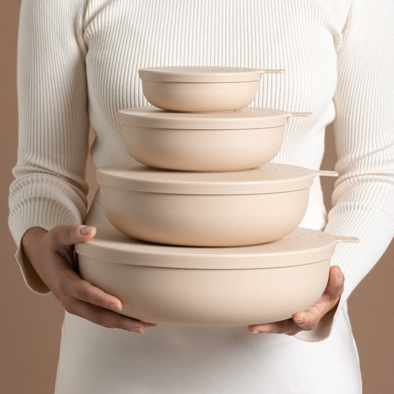Person holding a stack of beige plastic bowls with lids against a neutral background