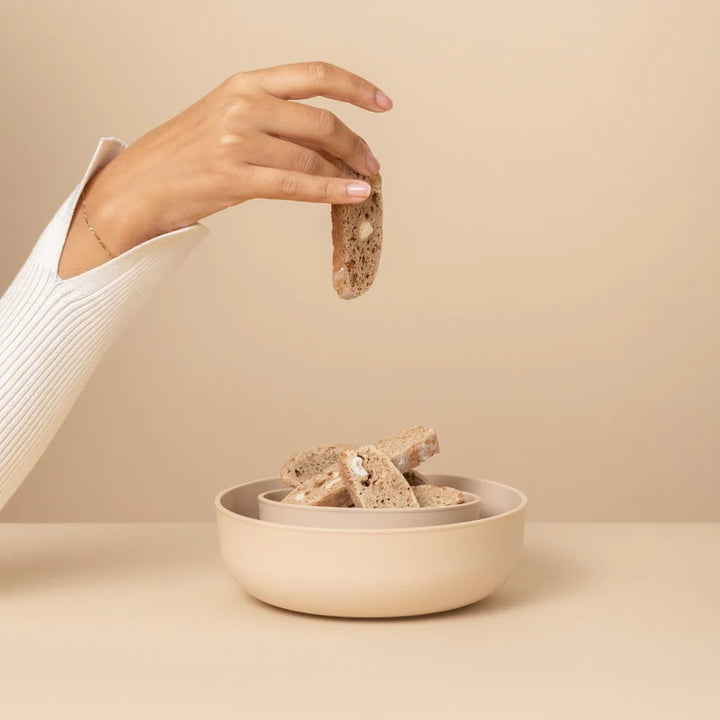 Hand placing a piece of biscotti into a bowl on a beige background