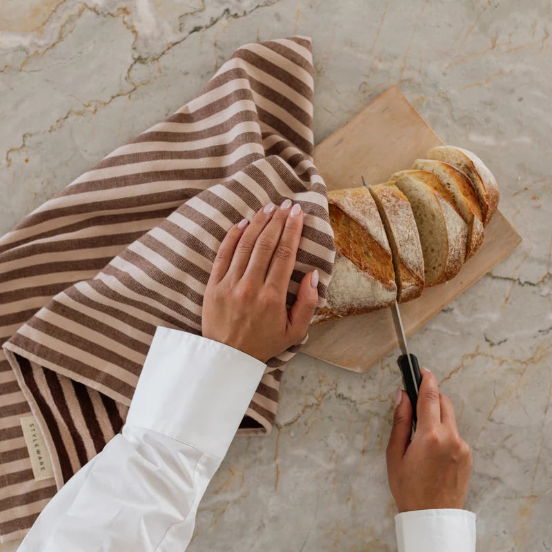 Person slicing bread on a wooden board with a striped towel on a marble countertop