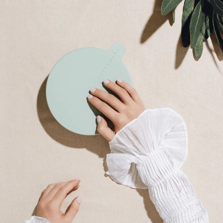 Hand holding a light green bowl with lid on a beige surface with a plant shadow.