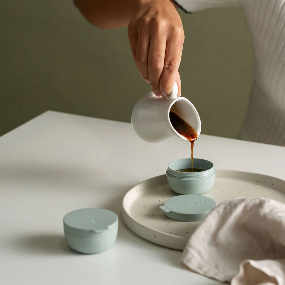 Person pouring sauce from a white cup into a small green container on a light surface.